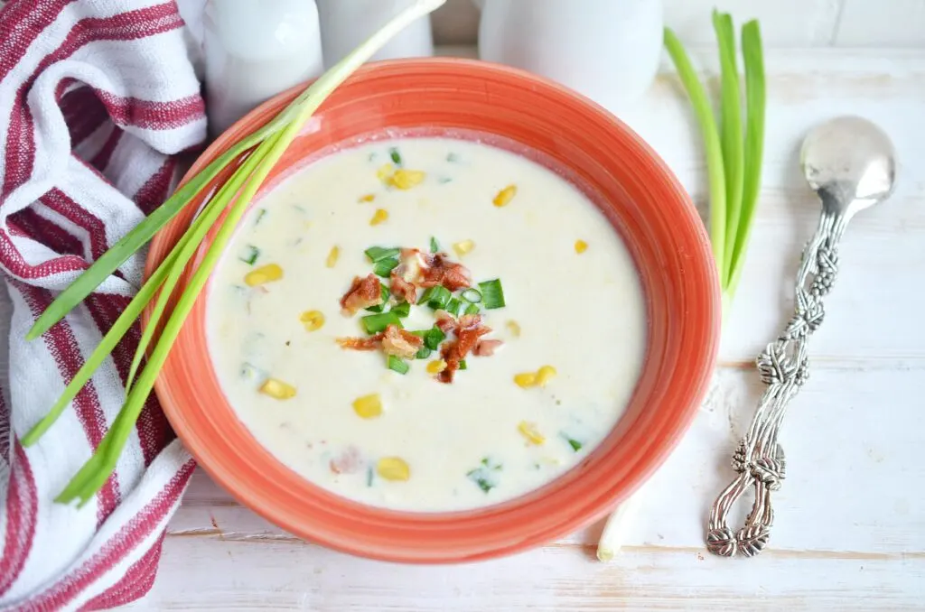 corn chowder in a red orange bowl with an onion and spoon and towel