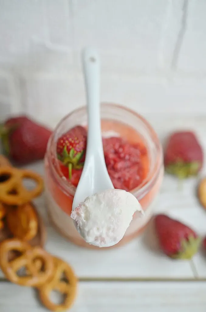Closeup of dessert in a spoon over layered strawberry pretzel salad in a jar with pretzels and strawberries to the side