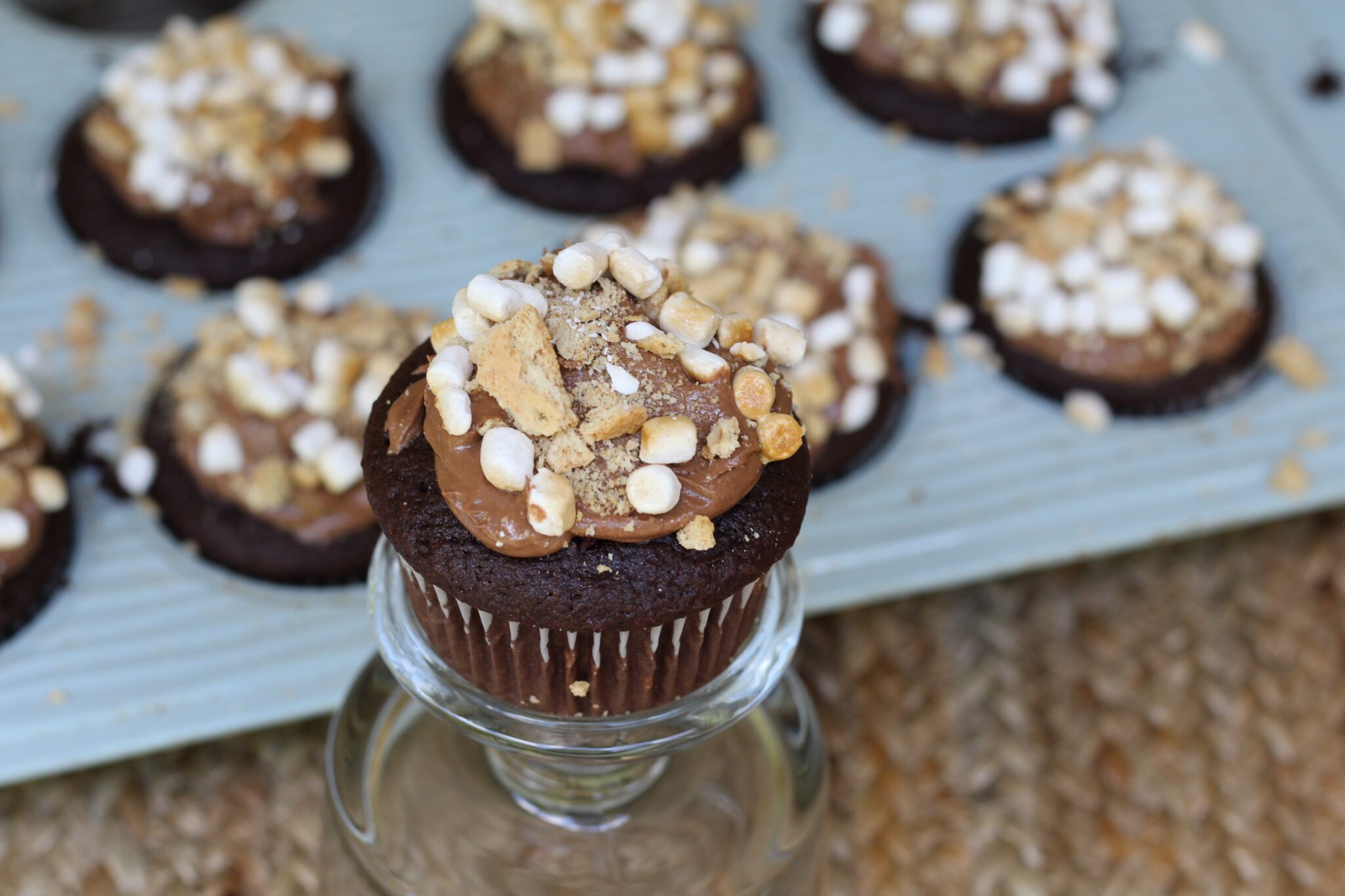 A s'mores cupcake in the foreground with a pan of cupcakes in the background, in a blue pan.